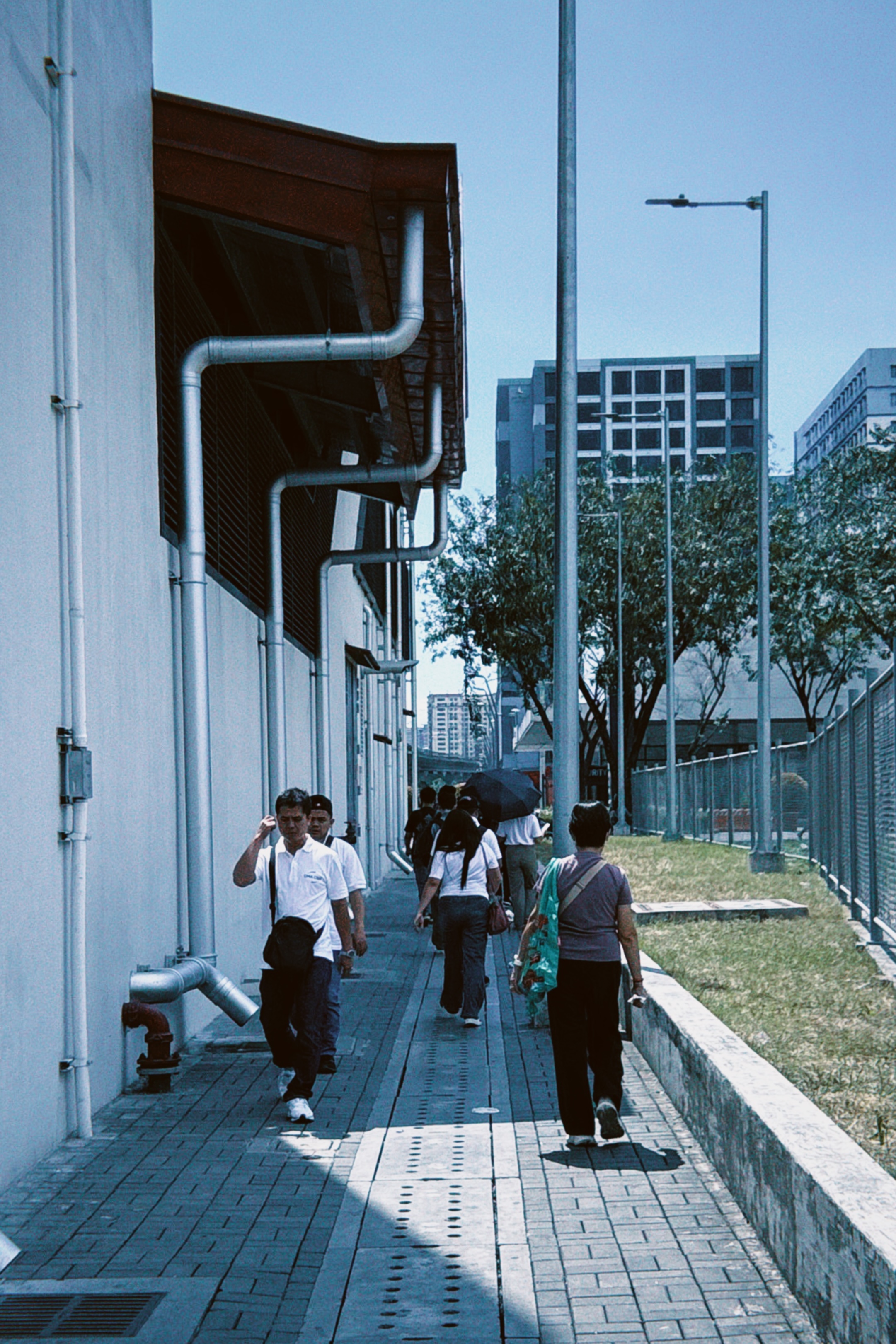 People walking along a footpath beside an LRT station building on a sunny day in Manila