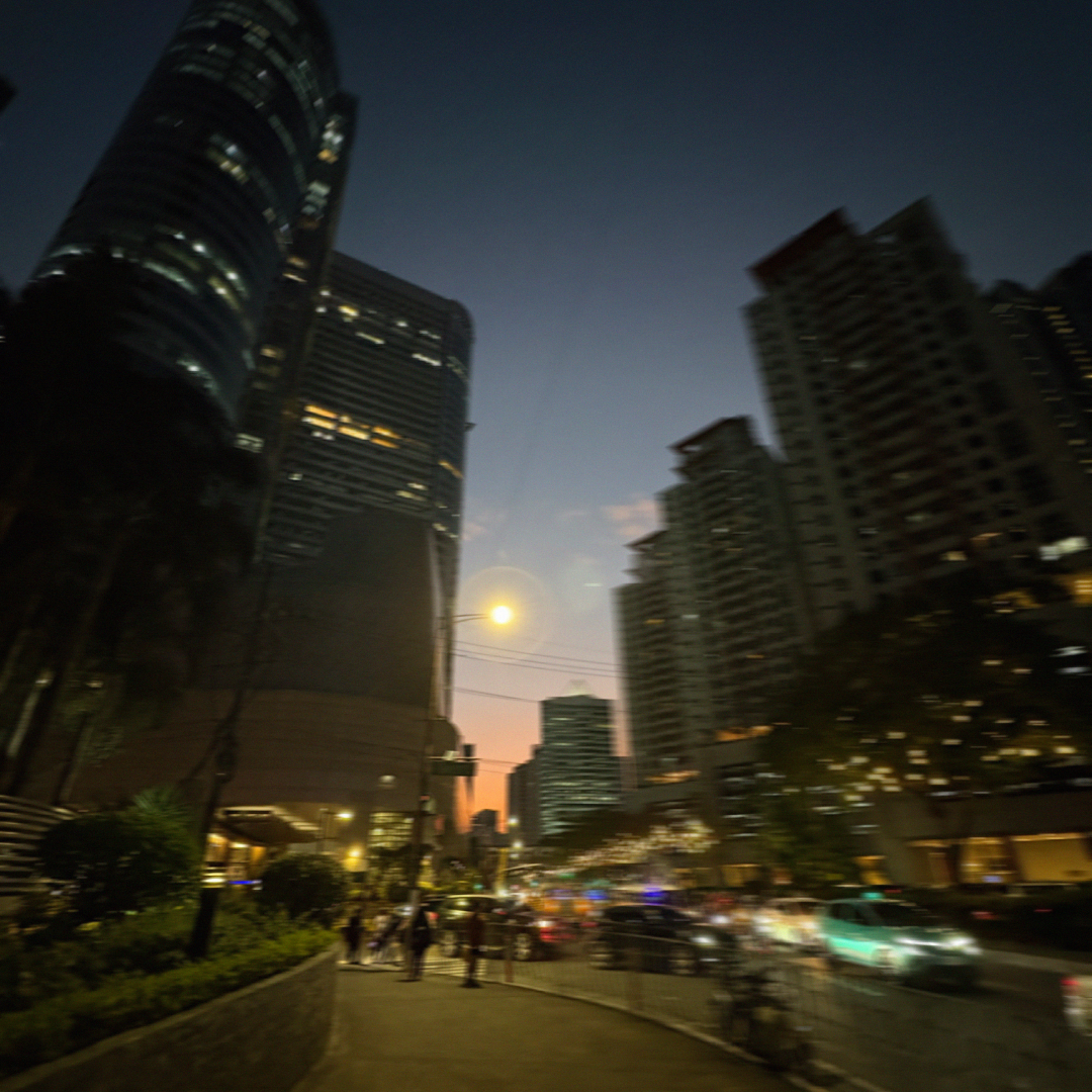 A nighttime street-level view of Makati with the RCBC Plaza tower on the left and blurred car lights and pedestrians along the road